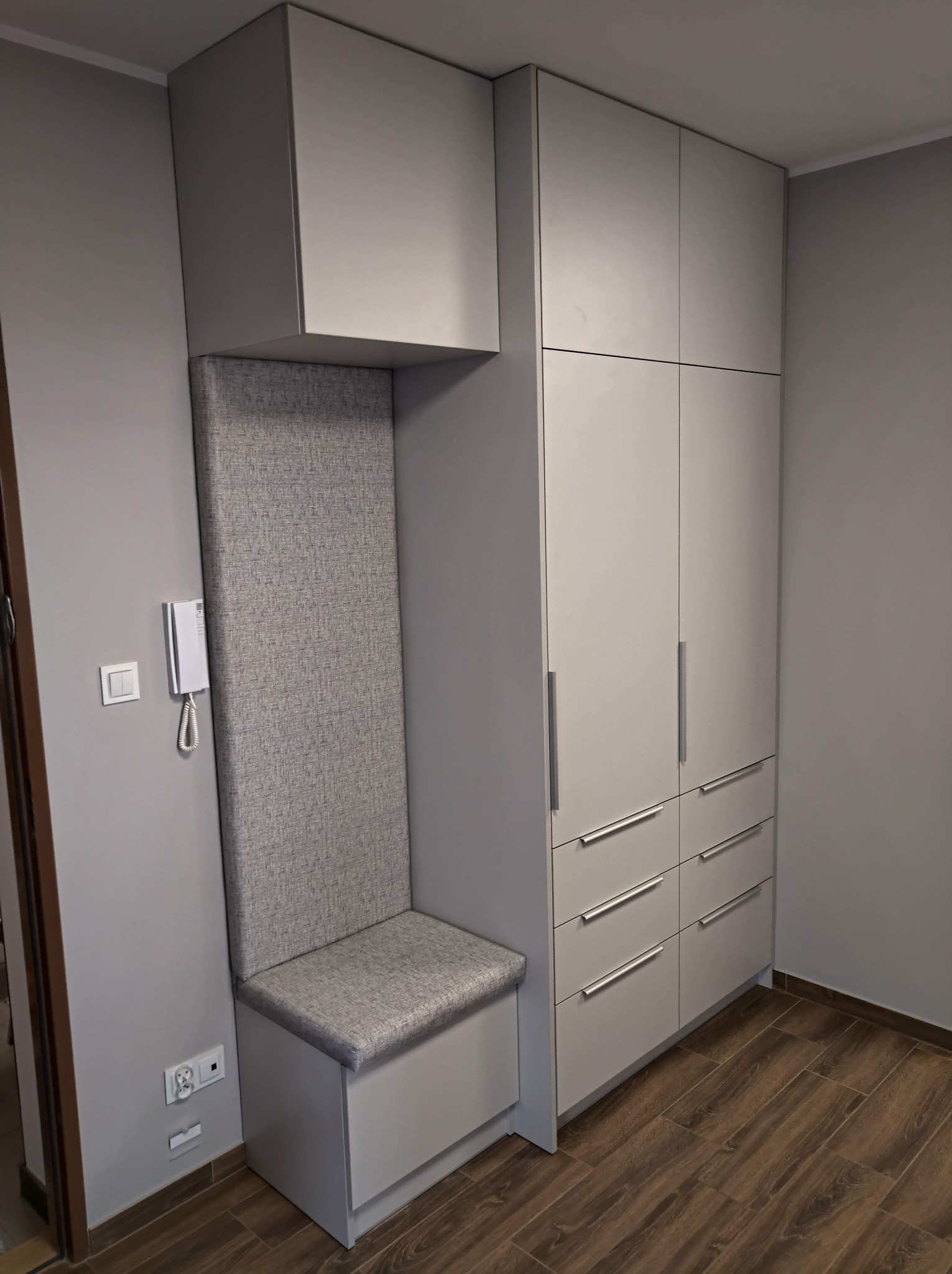 Szara zabudowa meblowa z siedziskiem i wysokim tapicerowanym oparciem w przedpokoju, obok drzwi wejściowych.
