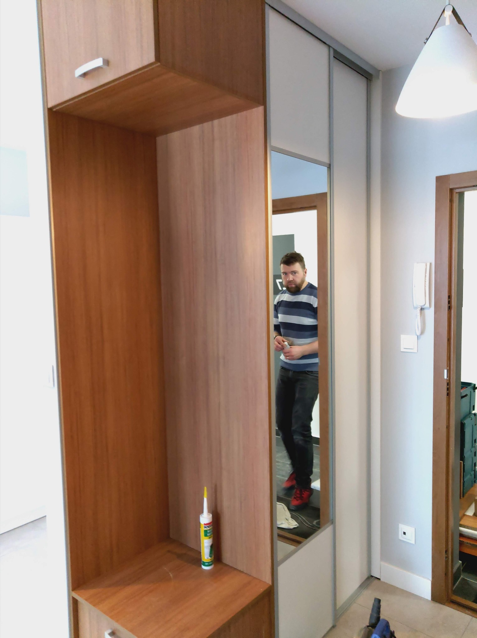 Wnętrze przedpokoju z zabudową meblową na wymiar w odcieniach brązu i bieli, widoczna tuba z klejem na półce, lustrzane drzwi przesuwne odbijające mężczyznę w paski.