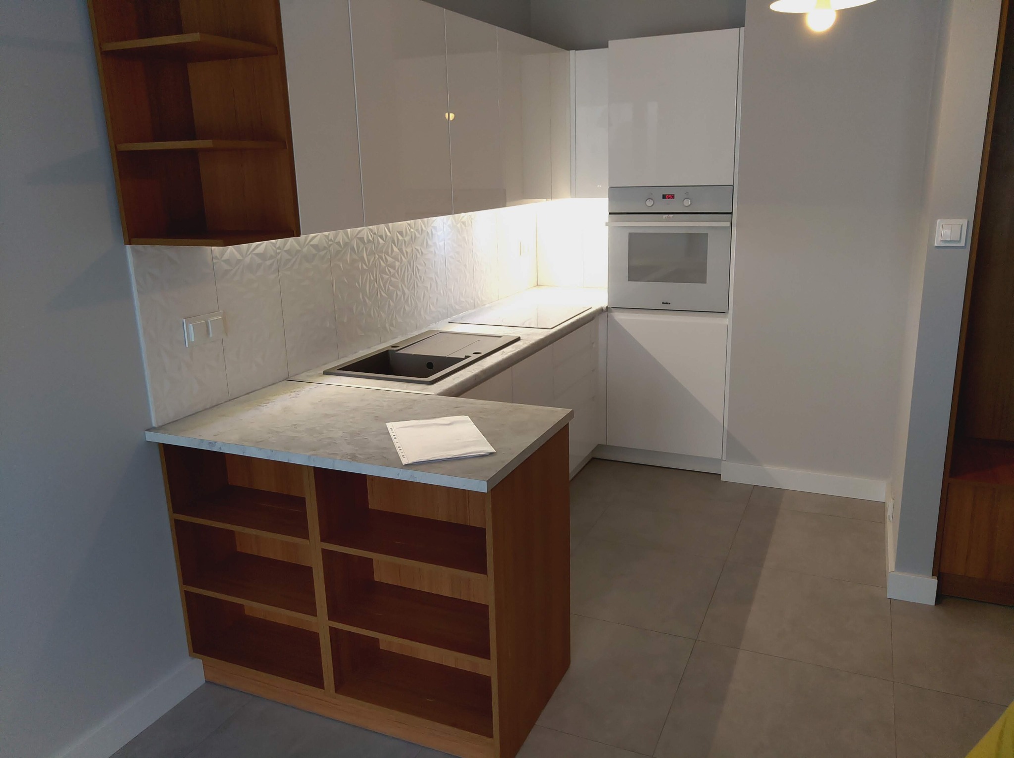 Nowoczesna, jasna kuchnia z białymi, lakierowanymi frontami szafek, podświetleniem LED pod szafkami wiszącymi i wyspą z drewnianymi półkami oraz blatem imitującym beton. Na blacie leży kartka papieru.