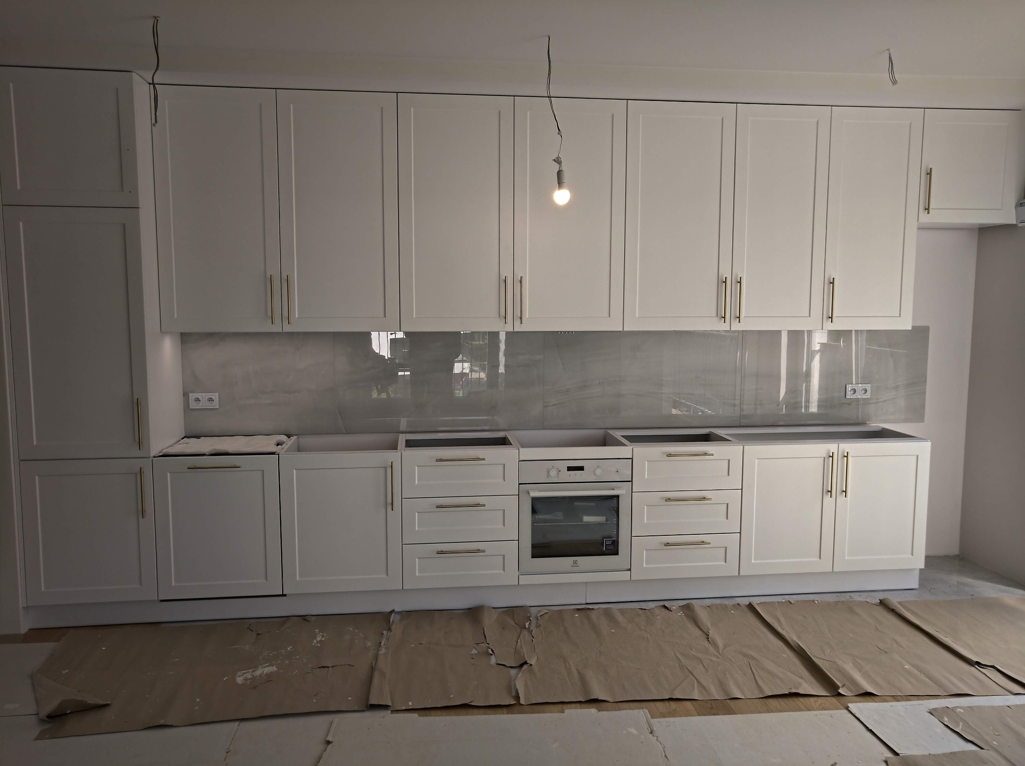 Montaż białych szafek kuchennych z marmurowym backsplashiem, widoczne zabezpieczenia podłogi i wisząca żarówka.