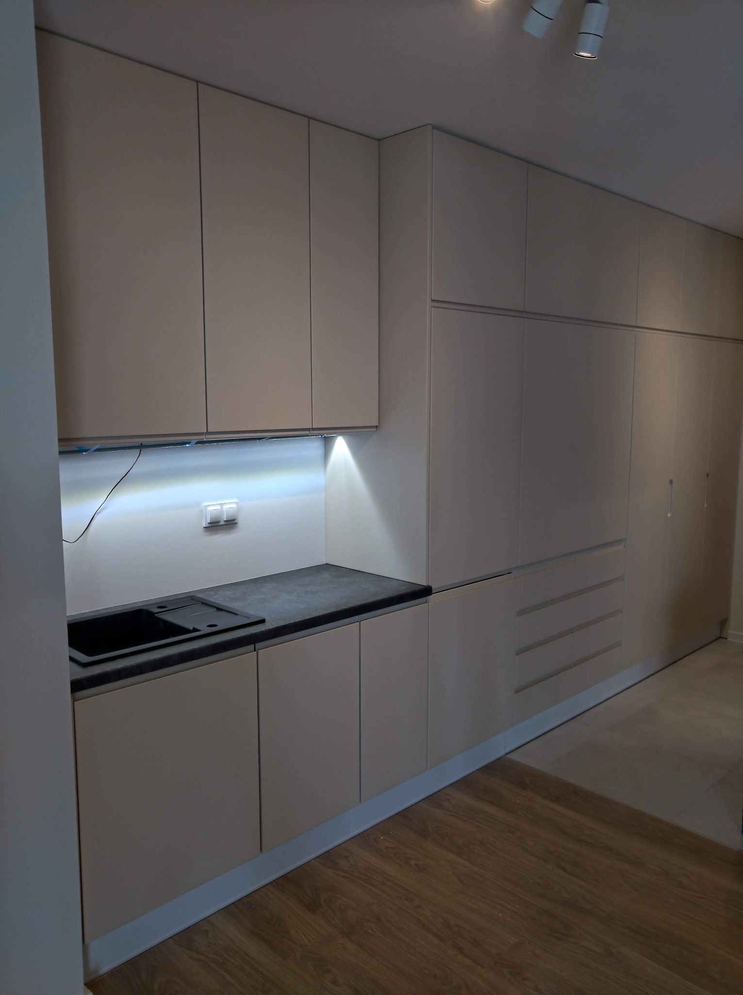 Beżowa zabudowa kuchenna z szafkami podwieszanymi i dolnymi, czarny zlew wpuszczany w blat, oświetlenie LED podszafkowe, drewniana podłoga.