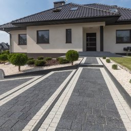 Eden Garden Firma Usługowa Damian Cichopek - Nowoczesny podjazd z ciemnoszarej kostki brukowej z jasnymi pasami, otoczony białym żwirem i niskimi krzewami, prowadzący do domu z ciemnym dachem.