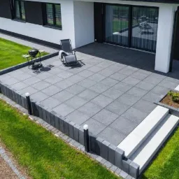 Nowoczesny taras z szarych płyt brukowych, obrzeża z ciemnych kostek, grill i leżak, trawnik, dom z dużymi oknami przesuwnymi.