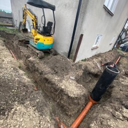 Eden Garden Firma Usługowa Damian Cichopek - Żółto-turkusowa mini koparka stoi na skraju wykopu, obok budynku. Pomarańczowe rury ułożone w wykopie, czarna rura karbowana w pierwszym planie. Mężczyzna w czarnej bluzie pracuje w wykopie.