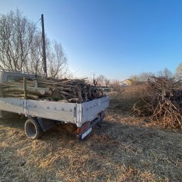 Eden Garden Firma Usługowa Damian Cichopek - Ciężarówka z platformą załadowana drewnem i gałęziami na zewnątrz, obok sterty suchych gałęzi, z widocznymi drzewami bez liści i fragmentem budynku w tle.