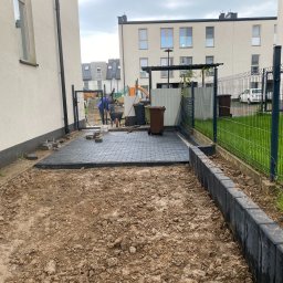 Eden Garden Firma Usługowa Damian Cichopek - Fragment układania szarej kostki brukowej na posesji, widoczni dwaj pracownicy, przygotowany teren z obrzeżami, w tle budynki mieszkalne i ogrodzenie panelowe.