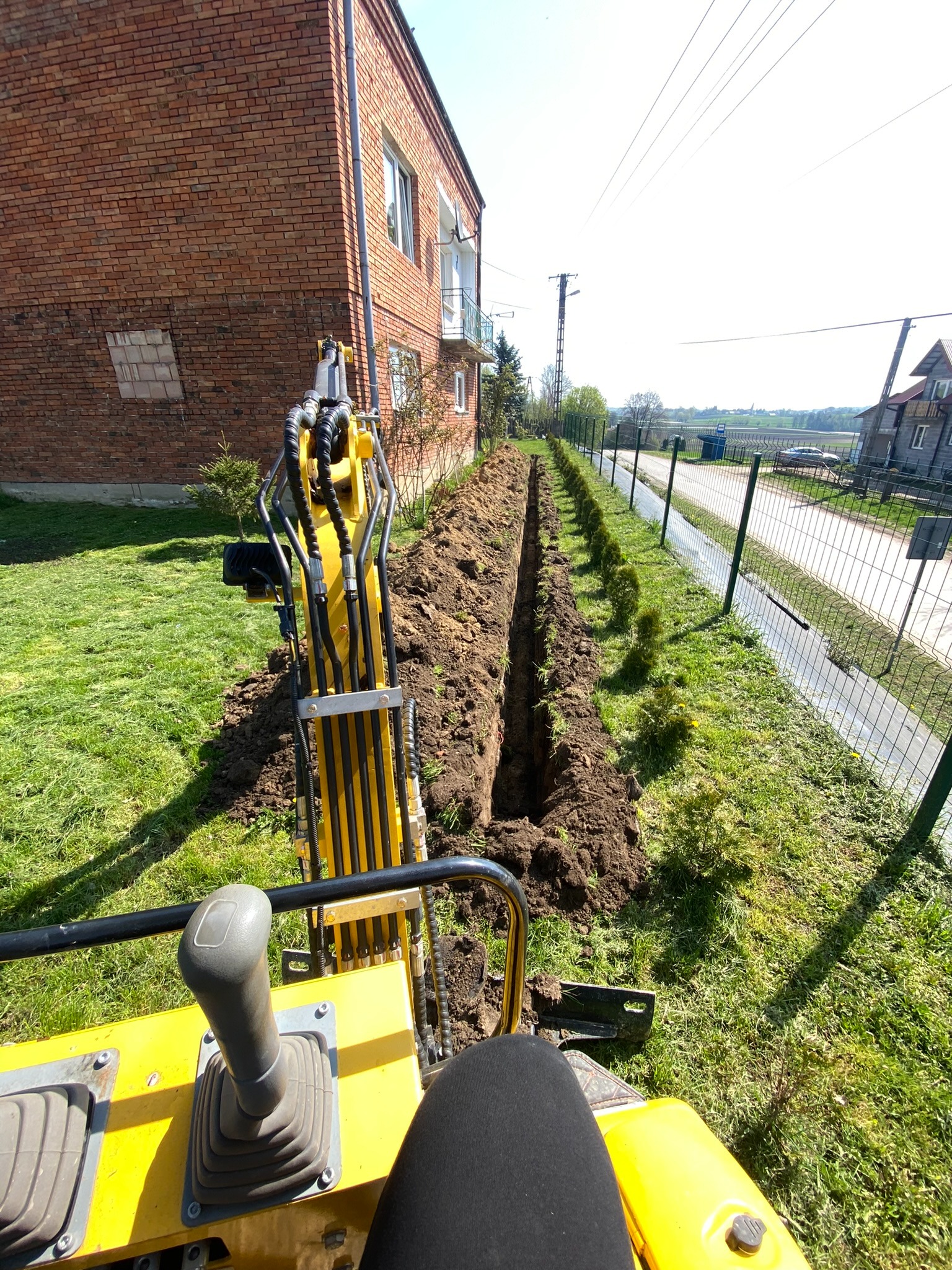 Widok z perspektywy operatora na żółtą mini koparkę, wykopany wąski rów wzdłuż ogrodzenia z siatki, budynek z czerwonej cegły w tle, słoneczny dzień.