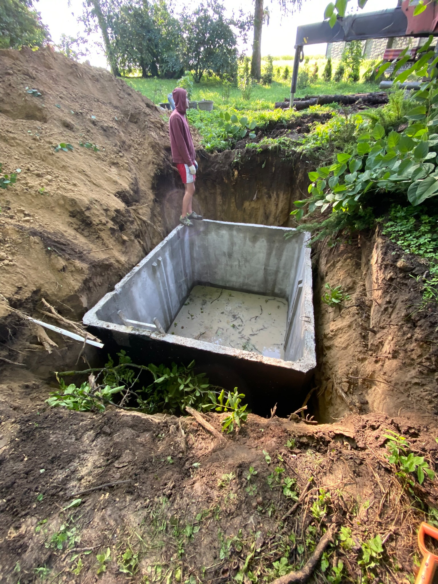 Wkopany w ziemię betonowy zbiornik, prawdopodobnie szambo, z osobą w bluzie z kapturem stojącą na jego krawędzi, otoczony wykopaną ziemią i zielenią.