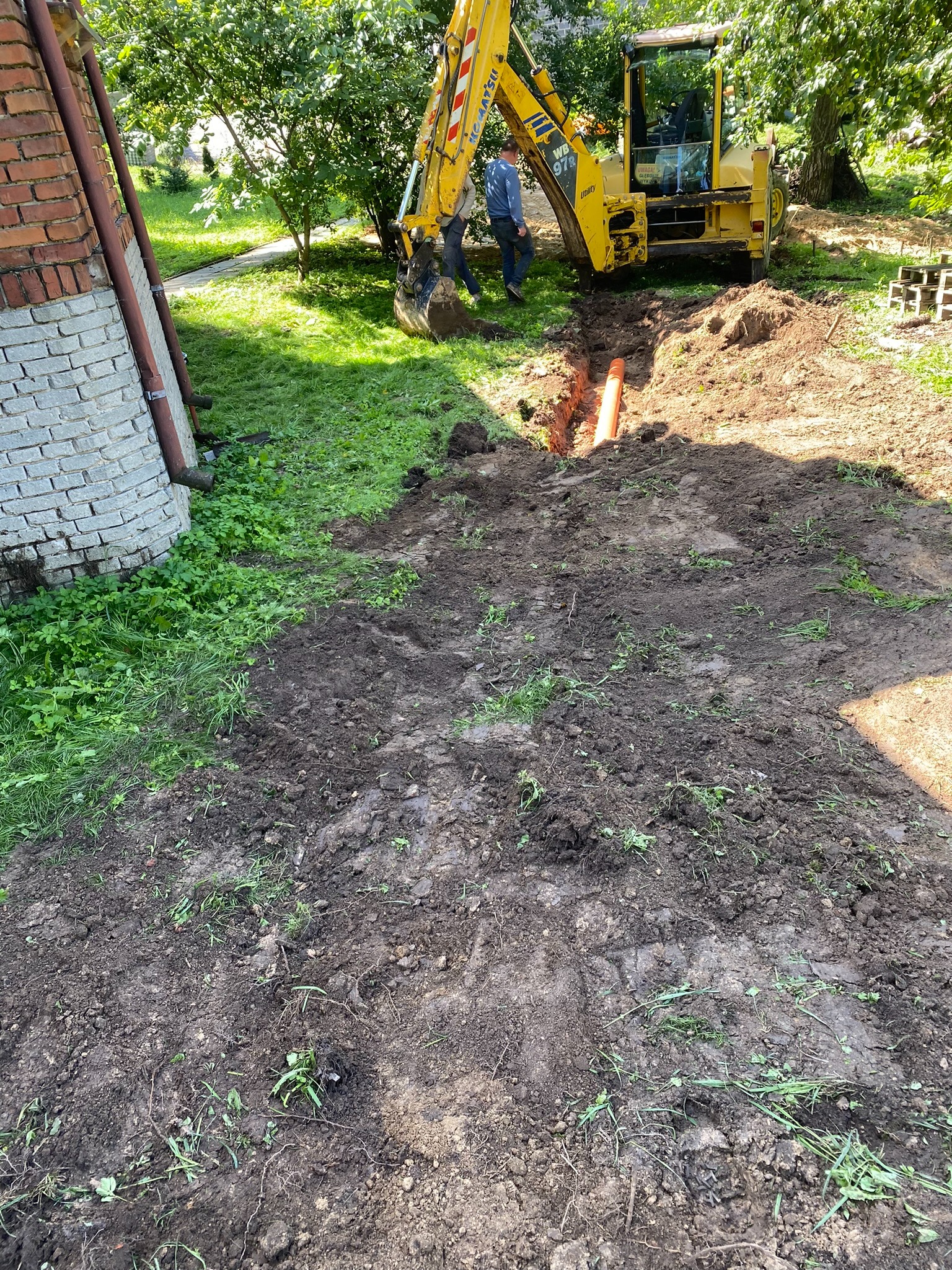 Żółta koparka wykonuje wykop pod rurę kanalizacyjną na trawiastym terenie obok budynku z cegły.
