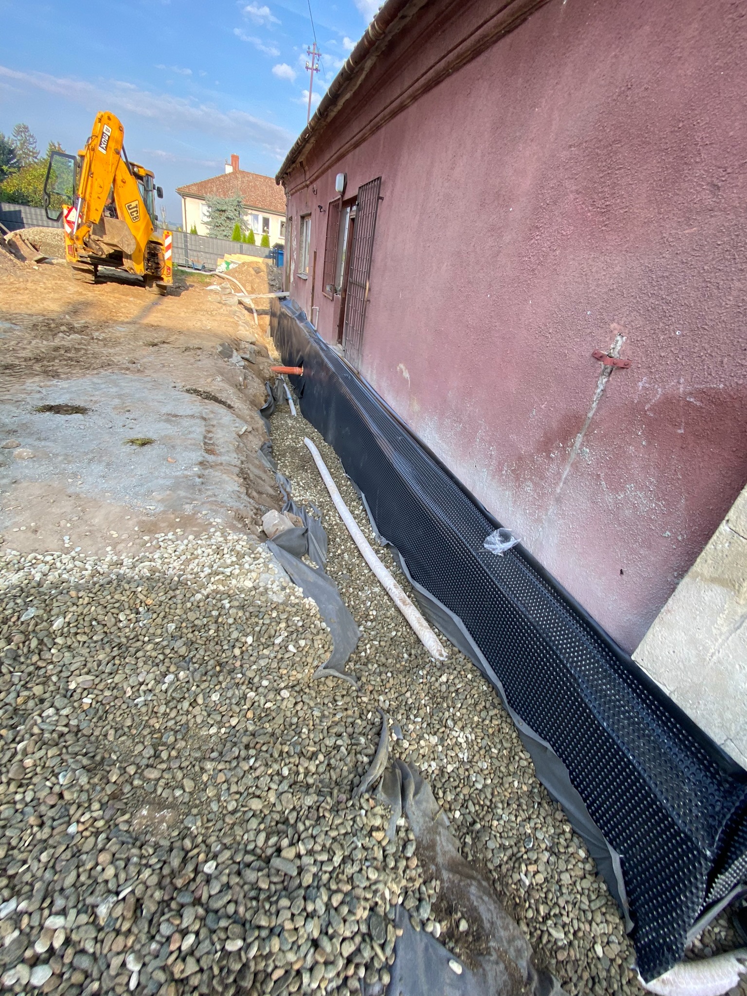 Prace ziemne przy budynku: wykop zabezpieczony folią kubełkową, rury drenażowe, zasypka z kamieni, w tle żółta koparka.