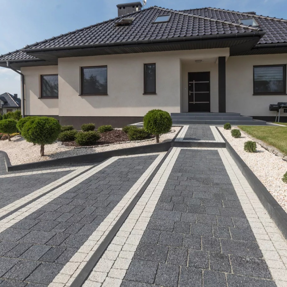 Nowoczesny podjazd z ciemnoszarej kostki brukowej z jasnymi pasami, otoczony białym żwirem i niskimi krzewami, prowadzący do domu z ciemnym dachem.