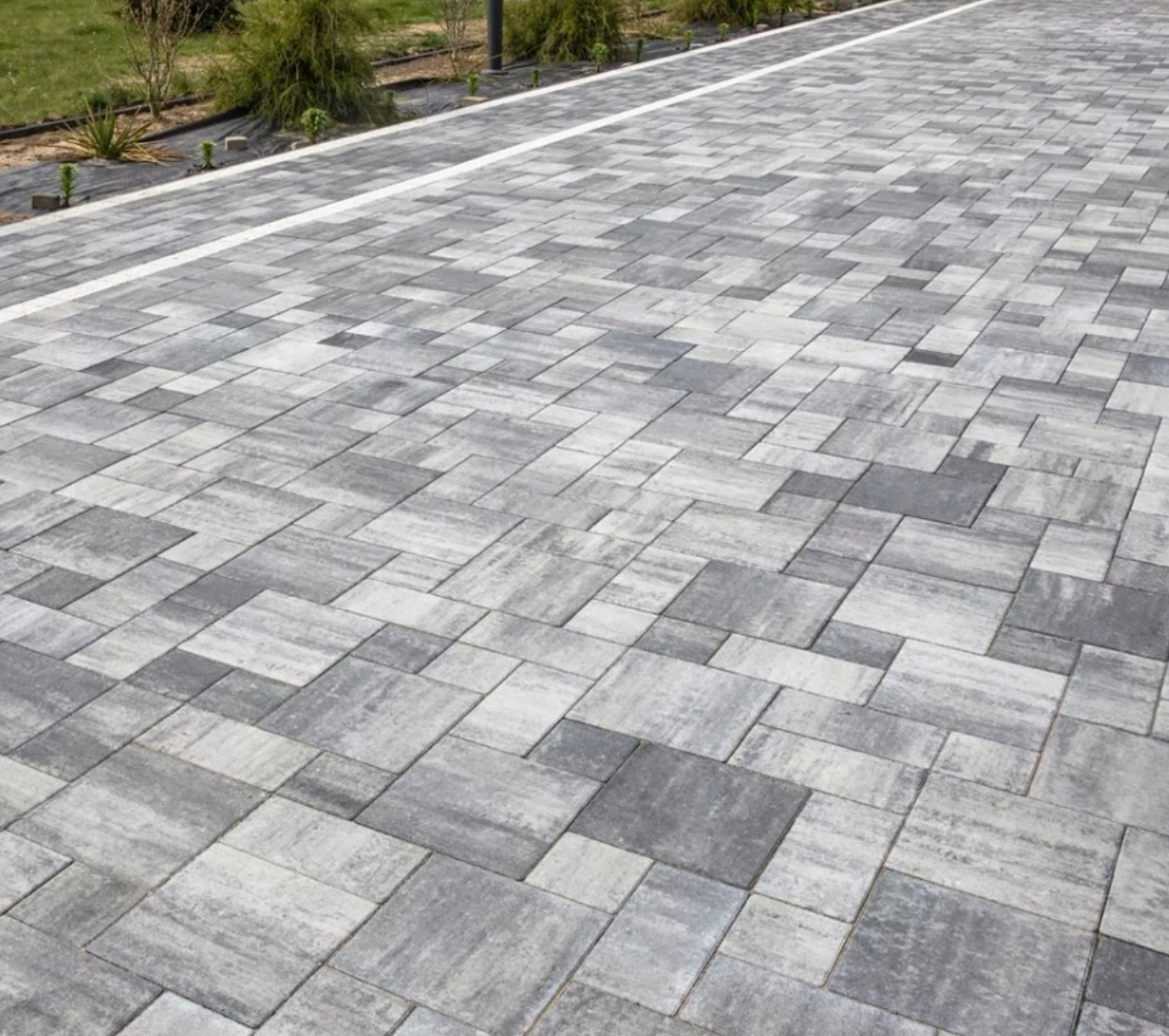 Ułożona nawierzchnia z szarej kostki brukowej o zróżnicowanej fakturze, oddzielona białym pasem od rabaty z niską zielenią i czarną agrowłókniną, widok z perspektywy.
