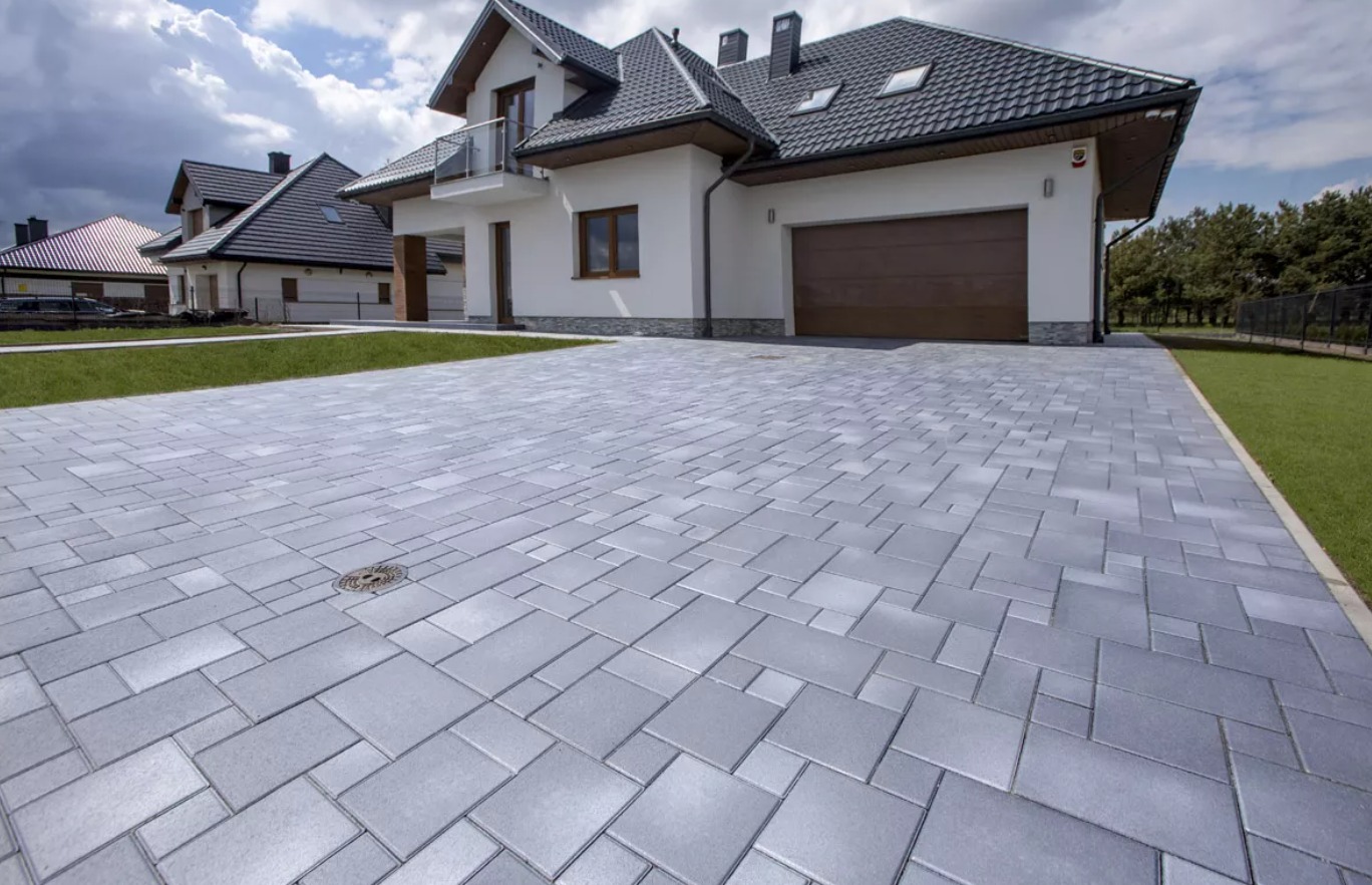 Nowoczesny podjazd z szarej kostki brukowej ułożonej we wzór, prowadzący do domu z garażem i balkonem. Widoczna studzienka kanalizacyjna wkomponowana w nawierzchnię.