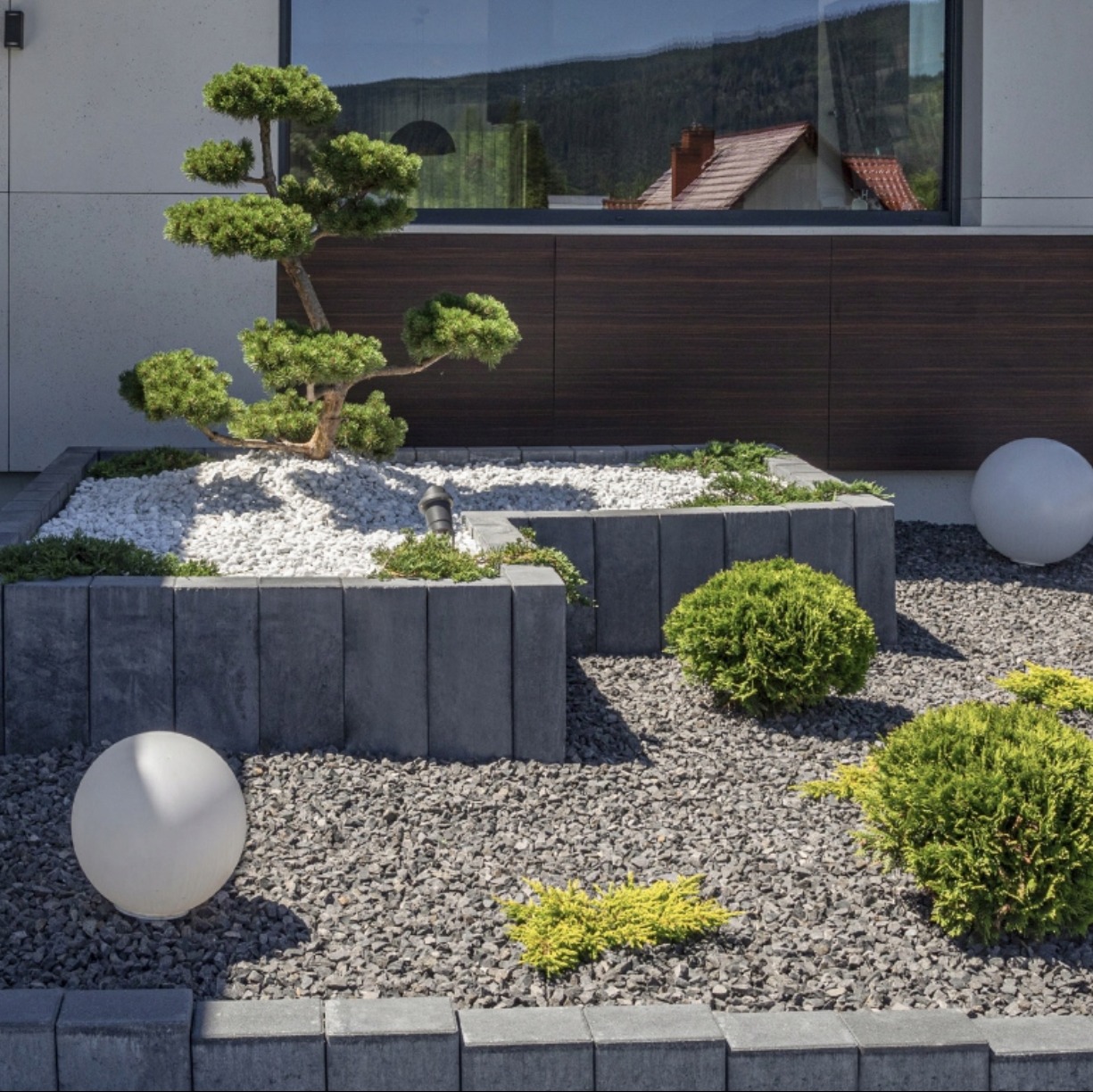 Nowoczesny ogród z podwyższonymi rabatami z szarej kostki, białym żwirem i formowanymi krzewami, oświetlony kulistymi lampami, z odbiciem domu w szybie okna.