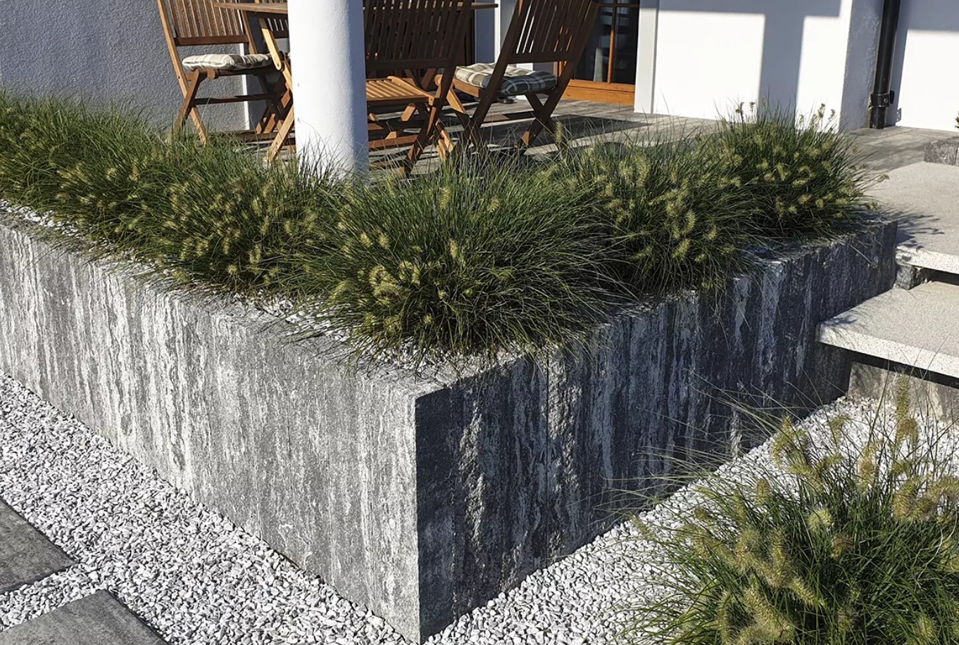 Podwyższona rabata z szarej kostki o nieregularnej fakturze, obsadzona ozdobnymi trawami, z białym żwirem u podstawy i elementami drewnianej architektury w tle.