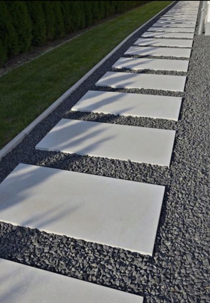 Prosta ścieżka z prostokątnych, jasnych płyt ułożonych na ciemnym żwirze, obok trawnika i żywopłotu. Cień drzewa pada na płyty.