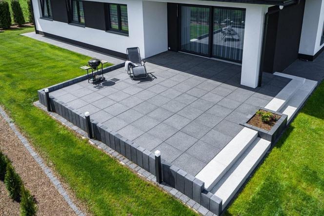 Nowoczesny taras z szarych płyt brukowych, obrzeża z ciemnych kostek, grill i leżak, trawnik, dom z dużymi oknami przesuwnymi.