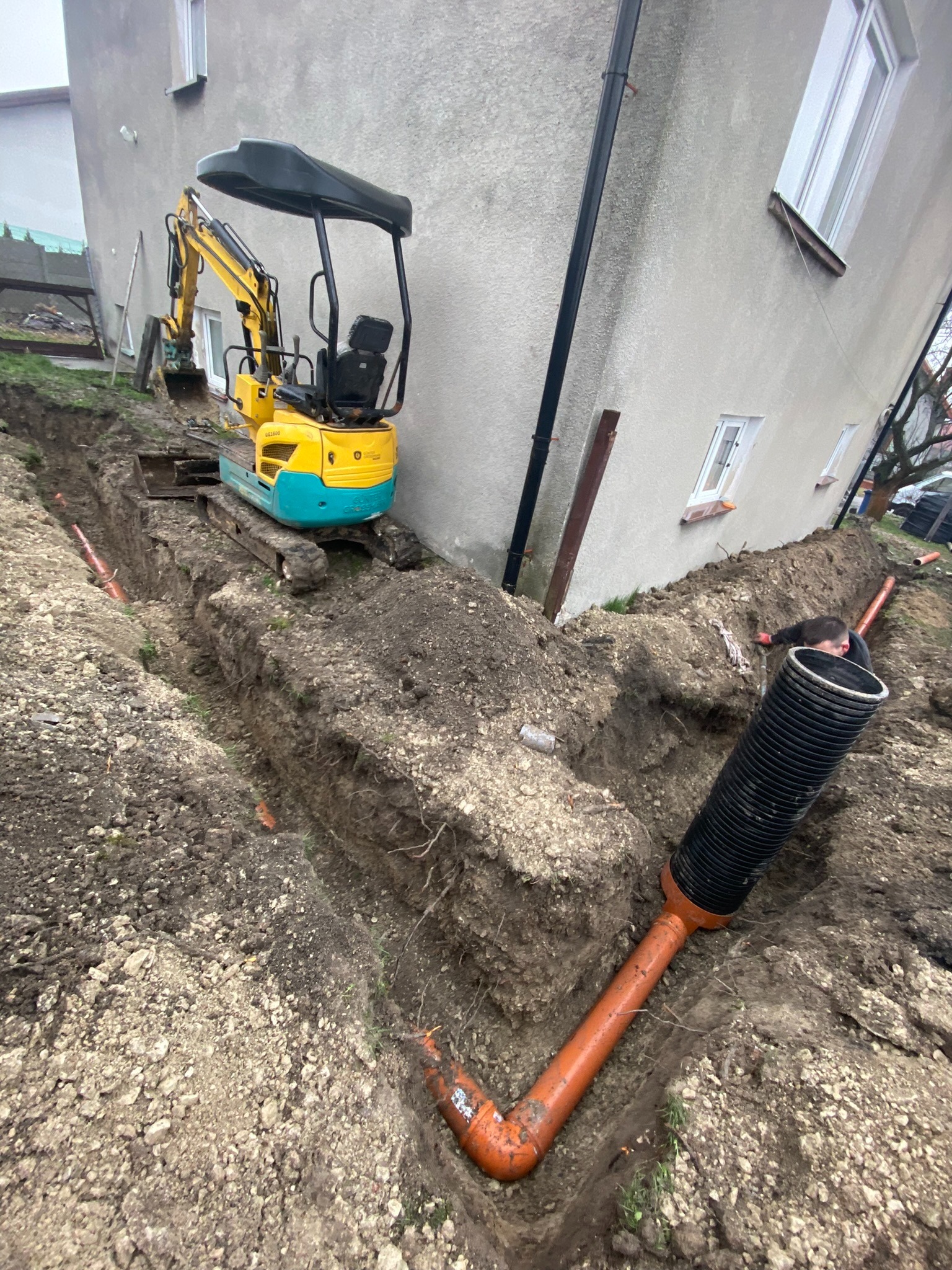 Żółto-turkusowa mini koparka stoi na skraju wykopu, obok budynku. Pomarańczowe rury ułożone w wykopie, czarna rura karbowana w pierwszym planie. Mężczyzna w czarnej bluzie pracuje w wykopie.