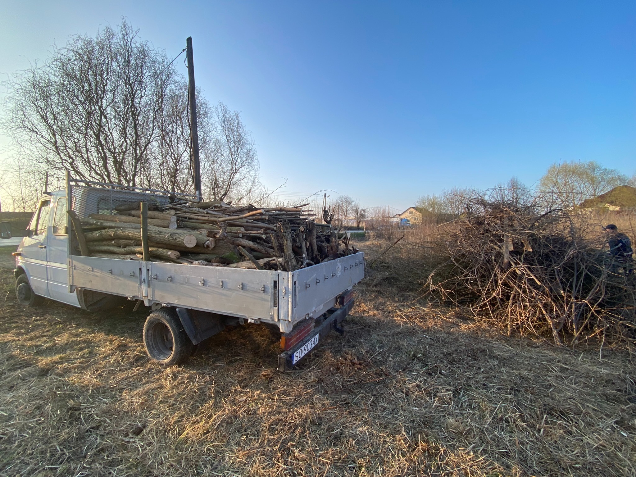 Ciężarówka z platformą załadowana drewnem i gałęziami na zewnątrz, obok sterty suchych gałęzi, z widocznymi drzewami bez liści i fragmentem budynku w tle.