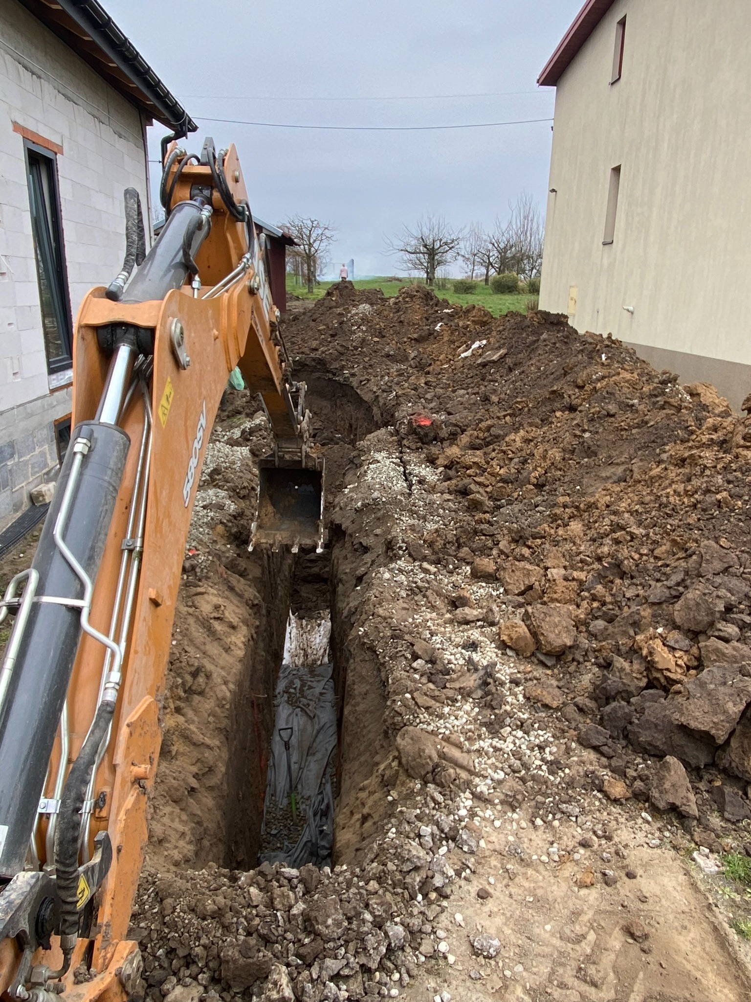 Pomarańczowa koparka CASE głębi wąski wykop w ziemi między dwoma budynkami, na dnie widoczne narzędzia i czarna folia, wykopana ziemia usypana wzdłuż wykopu, pochmurne niebo w tle.