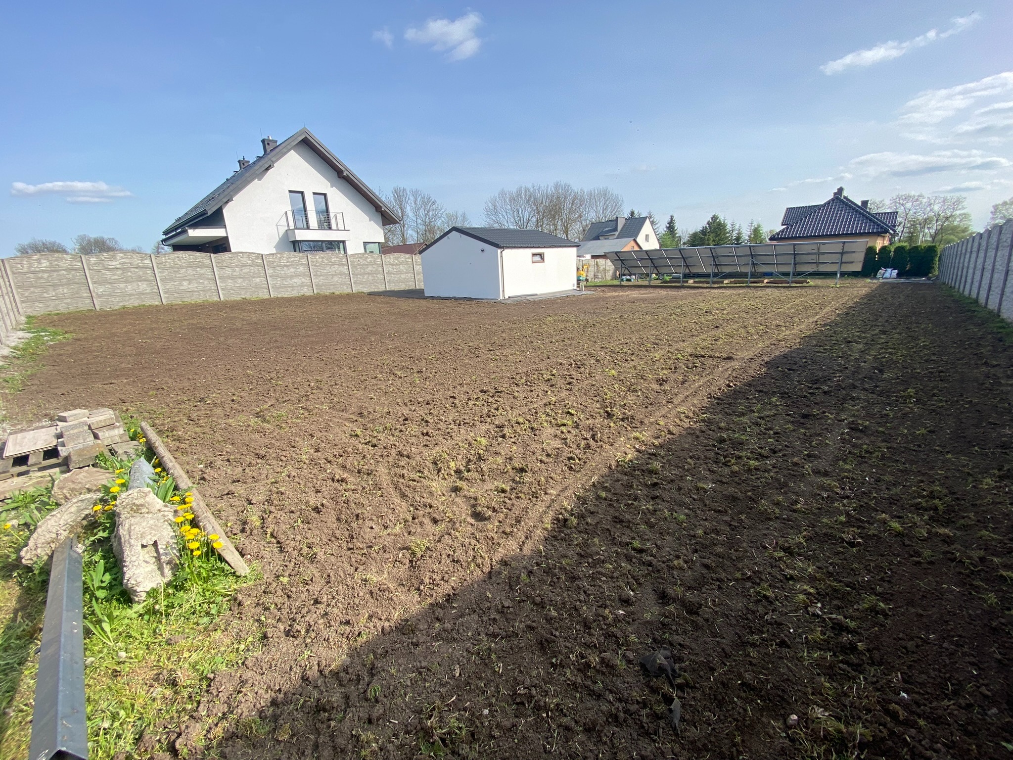 Przygotowany teren pod budowę z widocznymi śladami po maszynach, ogrodzony betonowym płotem, w tle dom z panelami słonecznymi i mały budynek gospodarczy.