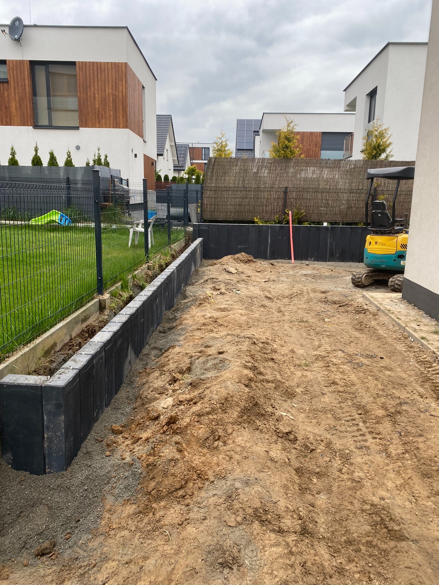Przygotowany teren pod budowę, widoczne czarne, betonowe bloczki krawężnikowe oddzielające trawnik od wykopu, mini koparka w tle, nowoczesne domy w zabudowie szeregowej w tle.