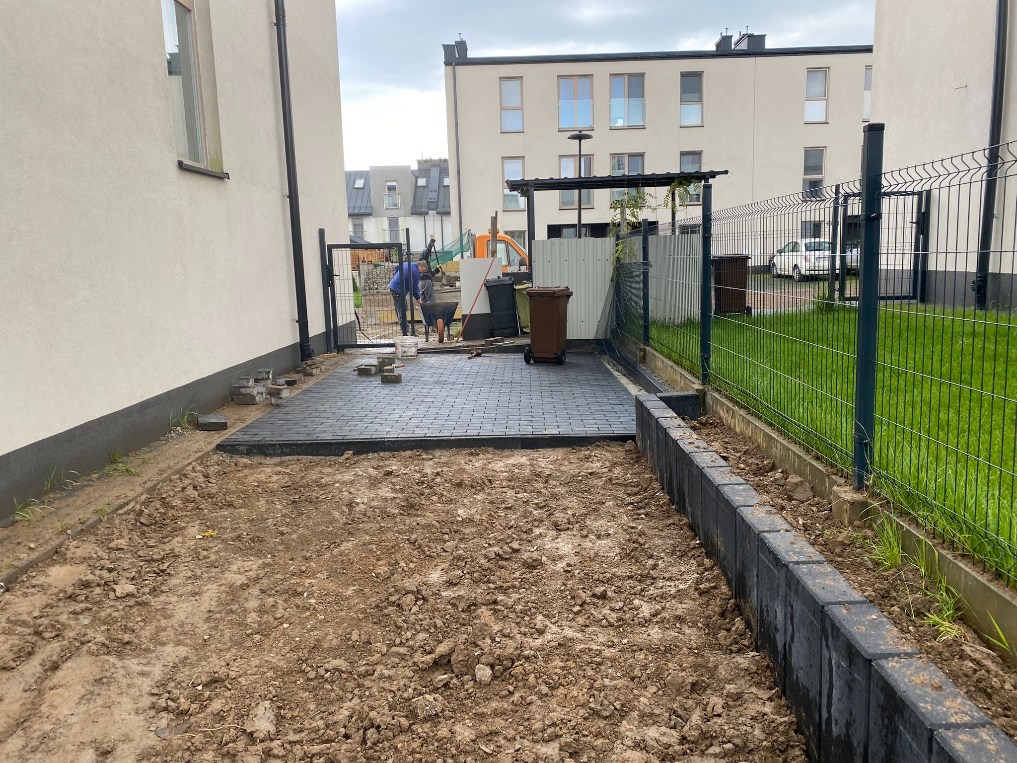 Fragment układania szarej kostki brukowej na posesji, widoczni dwaj pracownicy, przygotowany teren z obrzeżami, w tle budynki mieszkalne i ogrodzenie panelowe.