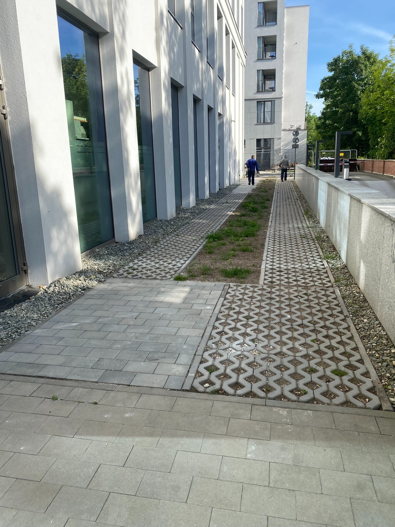 Ścieżka wyłożona różnymi rodzajami szarej kostki brukowej przed nowoczesnym budynkiem biurowym, z dwoma osobami idącymi w oddali.