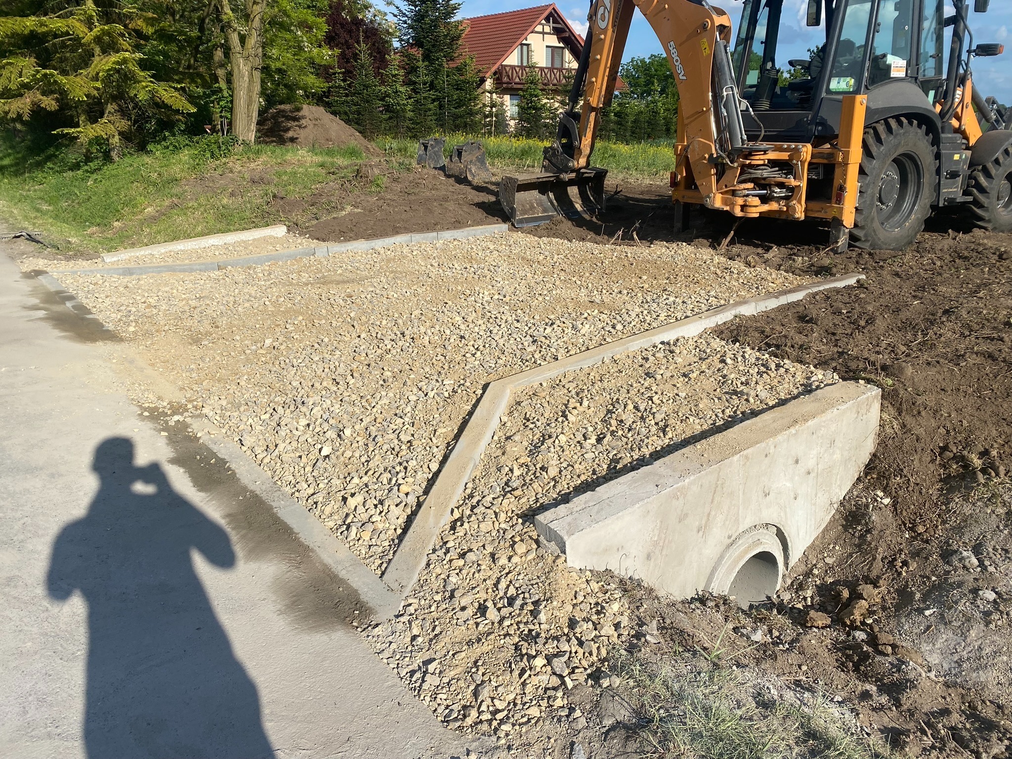 Żwirowe podłoże przygotowane pod nawierzchnię, obrzeża betonowe, rów odwadniający z przepustem oraz koparko-ładowarka Case w tle.