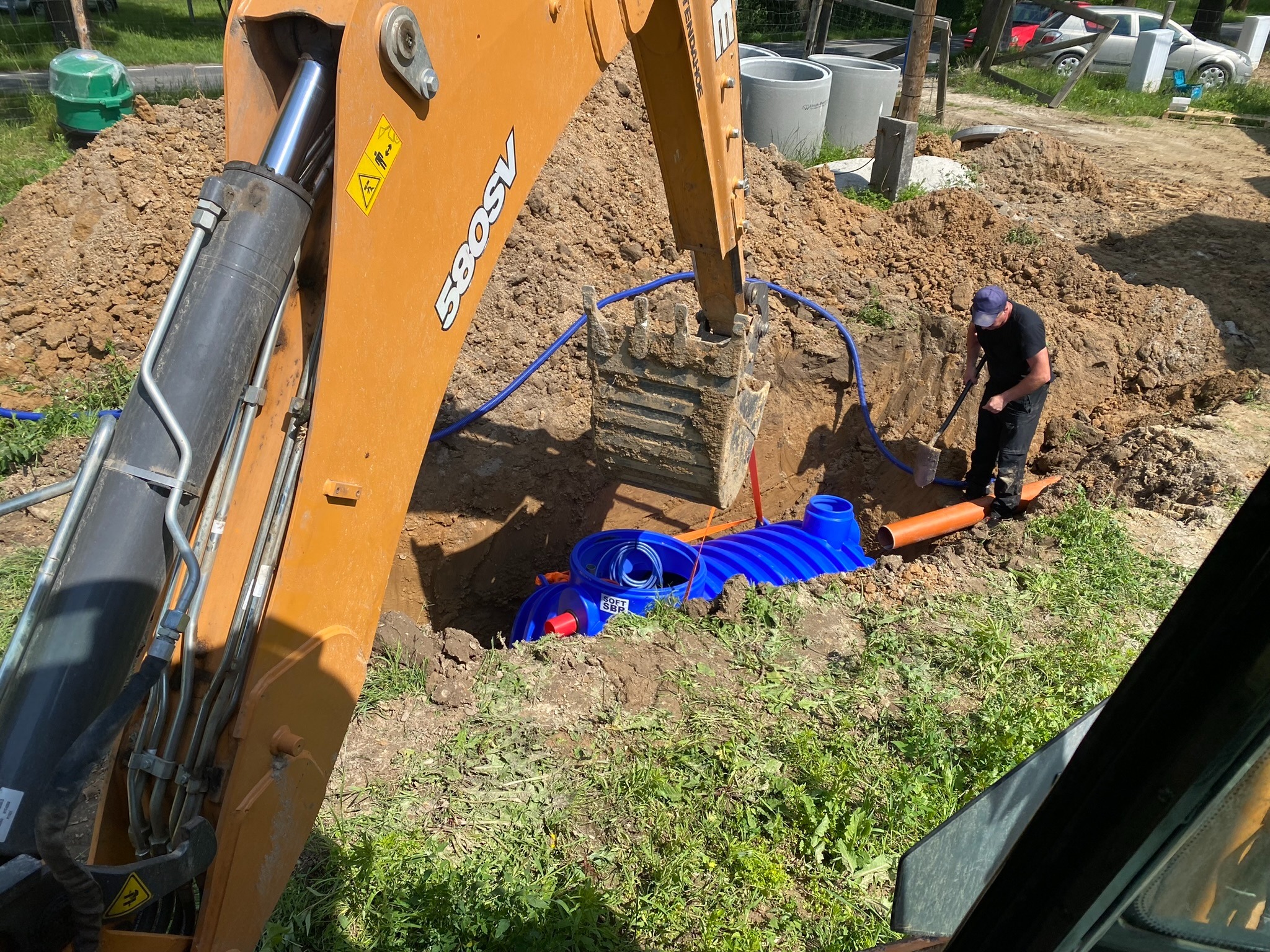 Pomarańczowa koparka CASE 580SV częściowo widoczna na pierwszym planie, zagłębia się łyżką w wykop, w którym umieszczono niebieski zbiornik z tworzywa, obok pracownik w czarnym ubraniu...