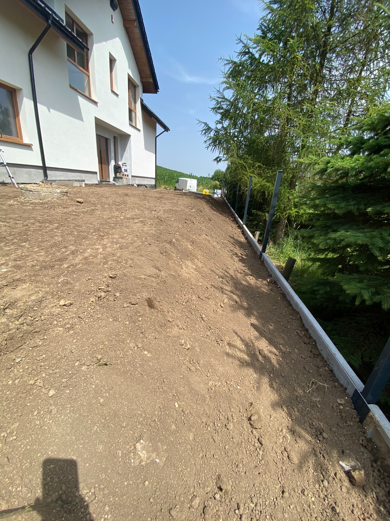 Przygotowany teren pod budowę ogrodzenia, widoczne słupki i obrzeża, w tle dom jednorodzinny i zieleń.