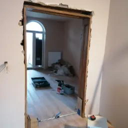 Przygotowanie do montażu ościeżnicy w surowym otworze drzwiowym z widocznym uszkodzeniem tynku, narzędzia na podłodze i drzwi balkonowe w tle.