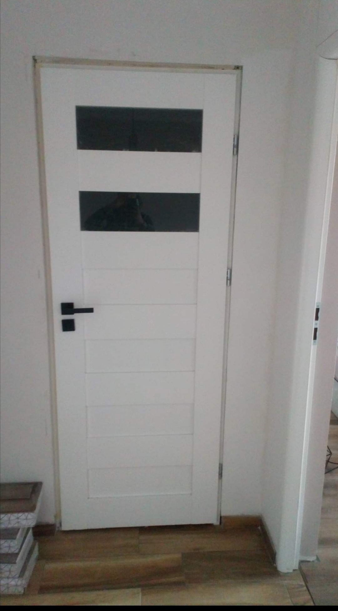 Białe drzwi wewnętrzne z dwoma poziomymi szybami, czarną klamką i ościeżnicą, widoczne na tle białej ściany i drewnopodobnej podłogi.