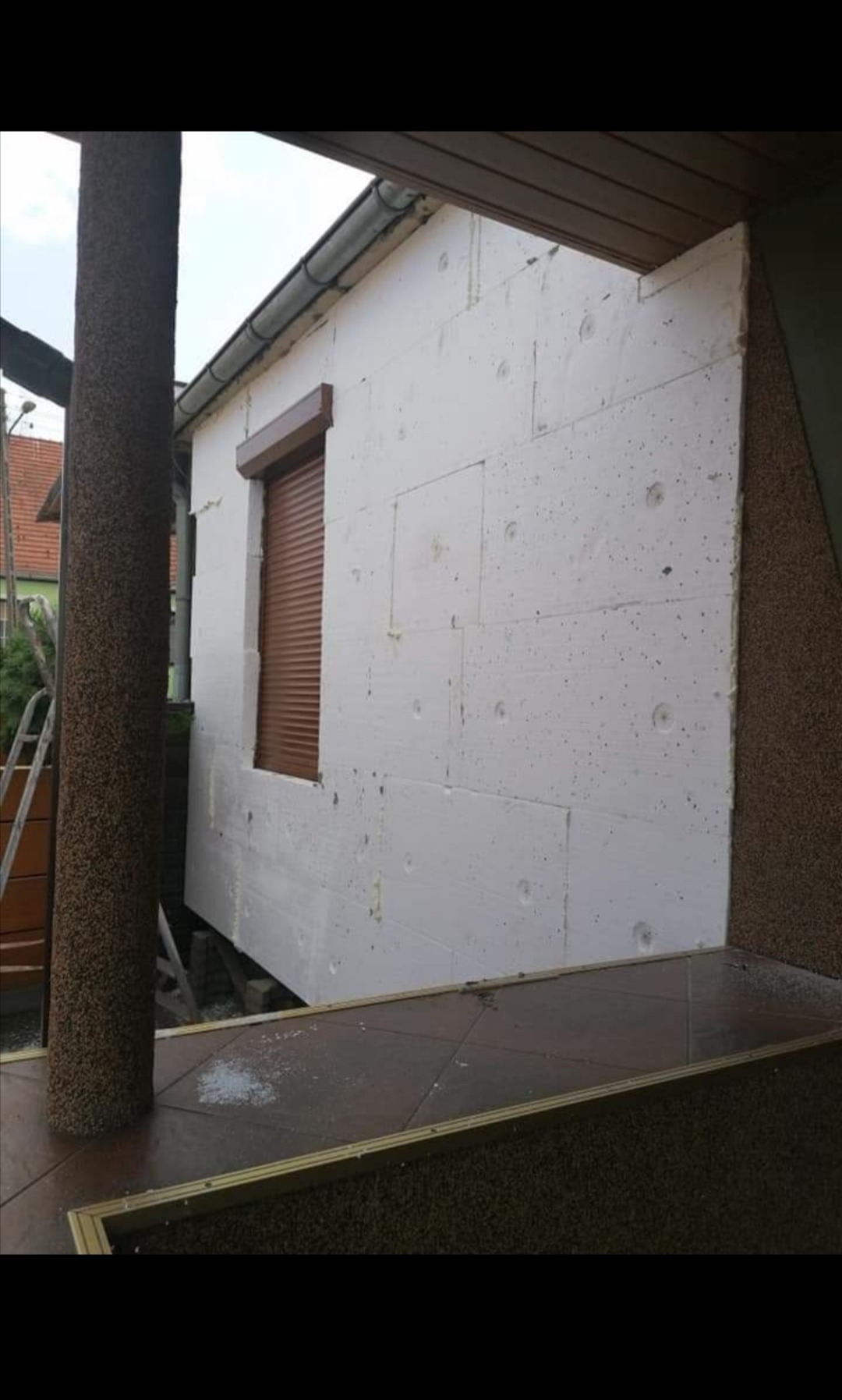 Ocieplenie budynku styropianem przed nałożeniem tynku, widoczne brązowe rolety okienne i fragment kolumny z fakturą kamienia.
