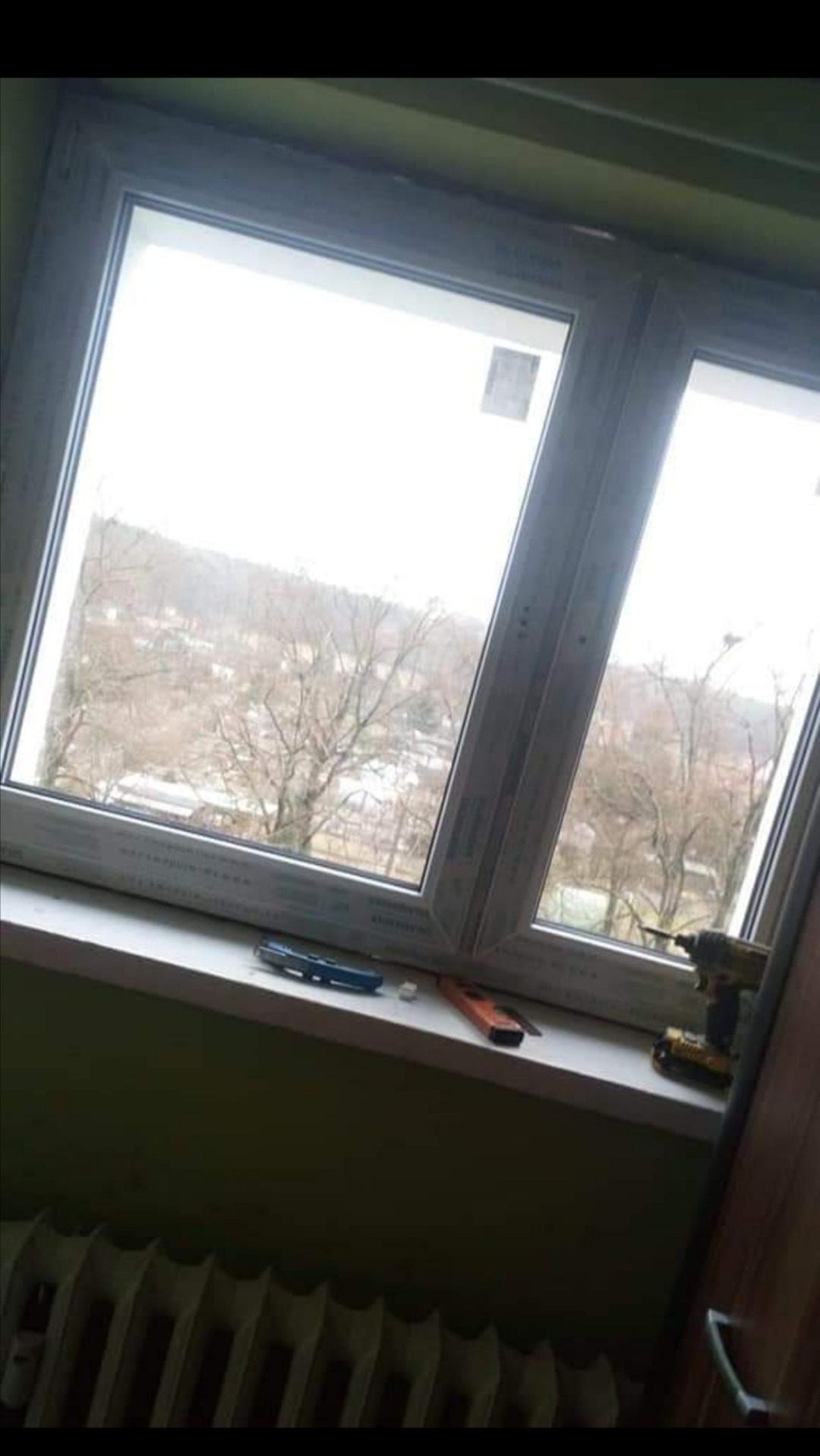 Nowe okno PCV z widokiem na miasto, na parapecie leżą narzędzia: wkrętarka, nożyk do tapet, a pod spodem stary grzejnik.