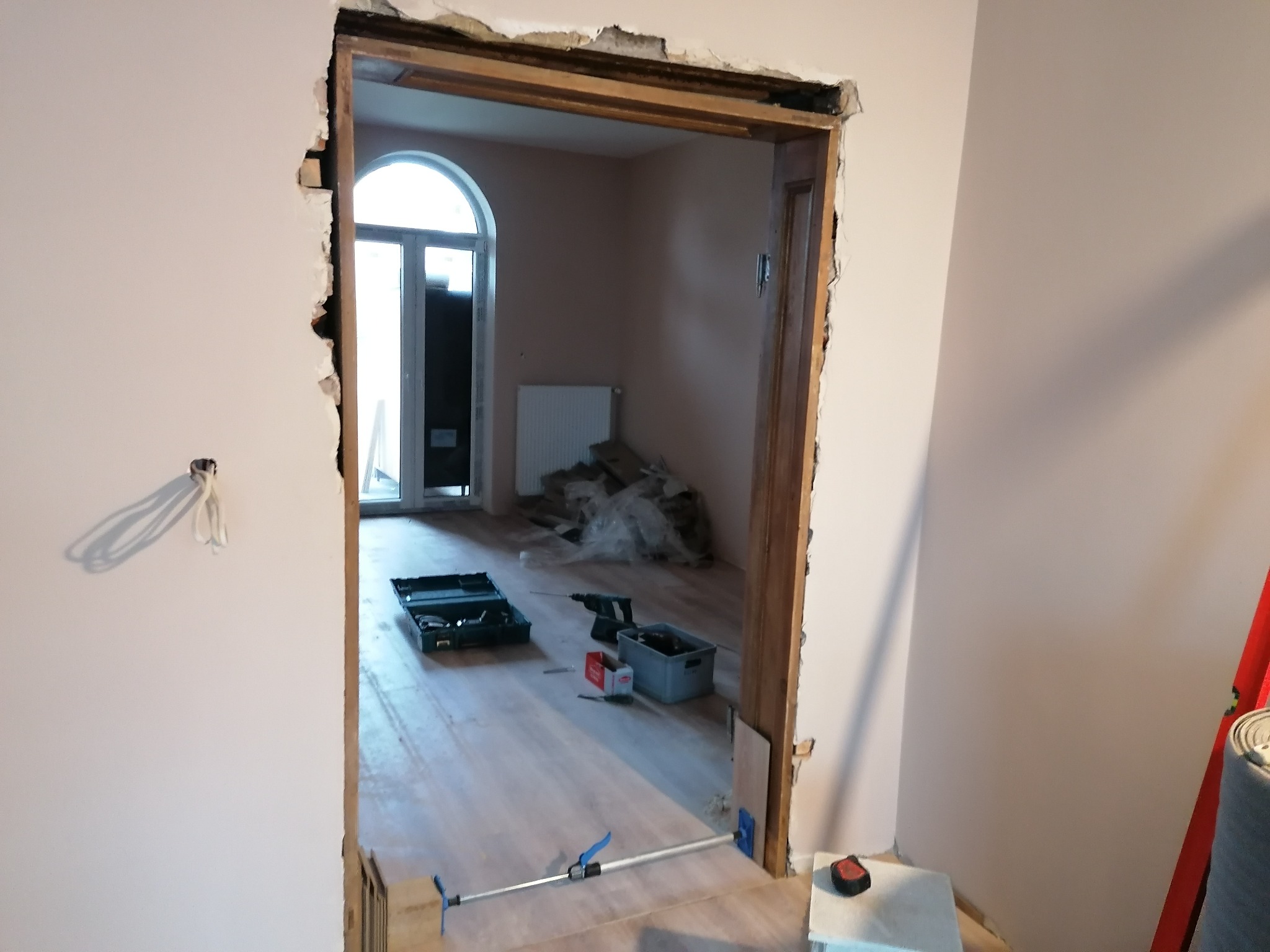 Przygotowanie do montażu ościeżnicy w surowym otworze drzwiowym z widocznym uszkodzeniem tynku, narzędzia na podłodze i drzwi balkonowe w tle.
