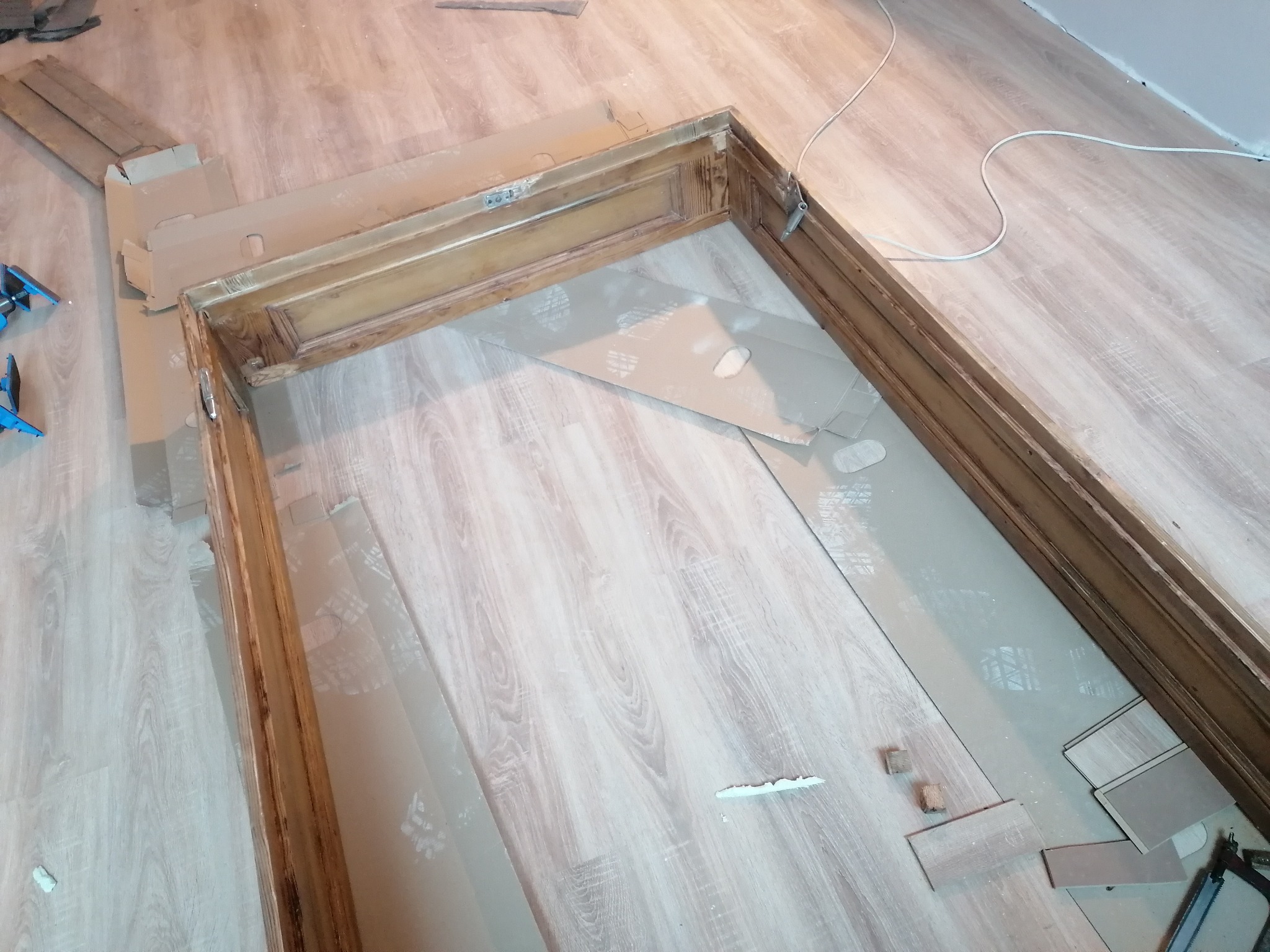 Demontaż ościeżnicy drzwiowej z widocznymi śladami po starym montażu, zabezpieczona kartonem podłoga z panelami w trakcie prac.