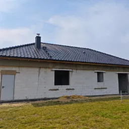 Dom w trakcie budowy z czarnym dachem z dachówki ceramicznej i widocznym kominem. Budynek z białych bloczków, okna i drzwi bez stolarki.