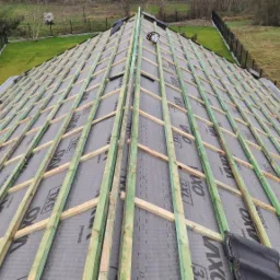 Widok z góry na konstrukcję dachu z łatami i kontrłatami na membranie dachowej, przygotowaną do krycia dachówką, widoczne narzędzie i elementy dachówki.