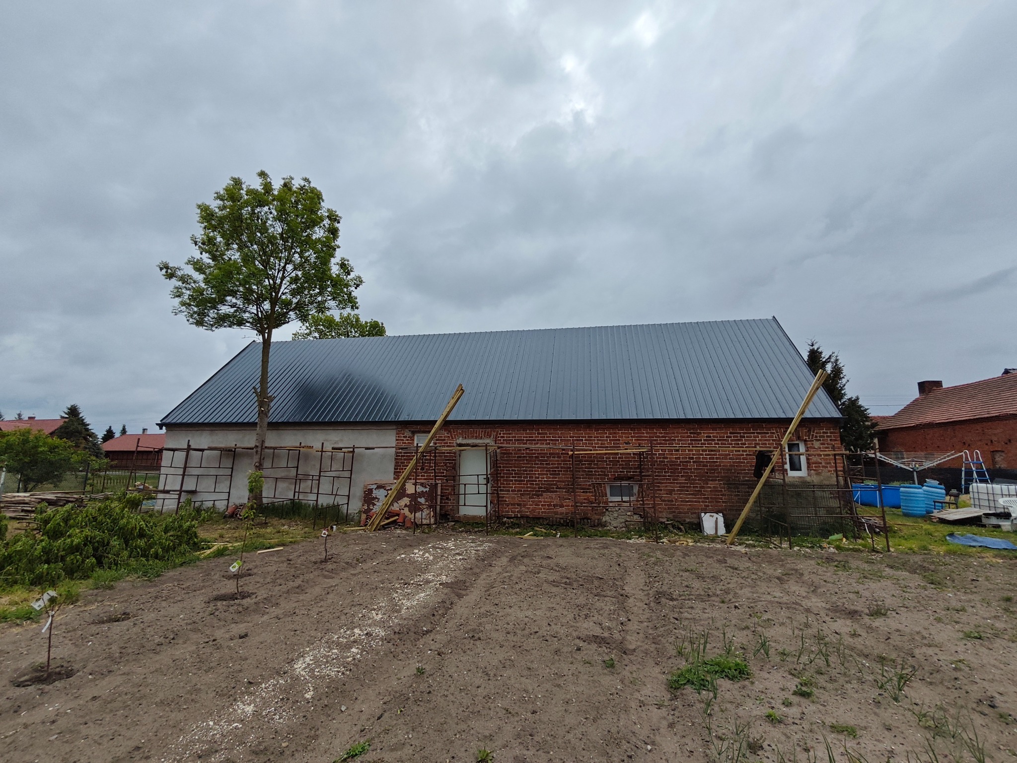 Nowy, blaszany dach na ceglanym budynku gospodarczym w trakcie budowy, z drewnianymi podporami i rusztowaniami. Na pierwszym planie świeżo zasadzone drzewka.