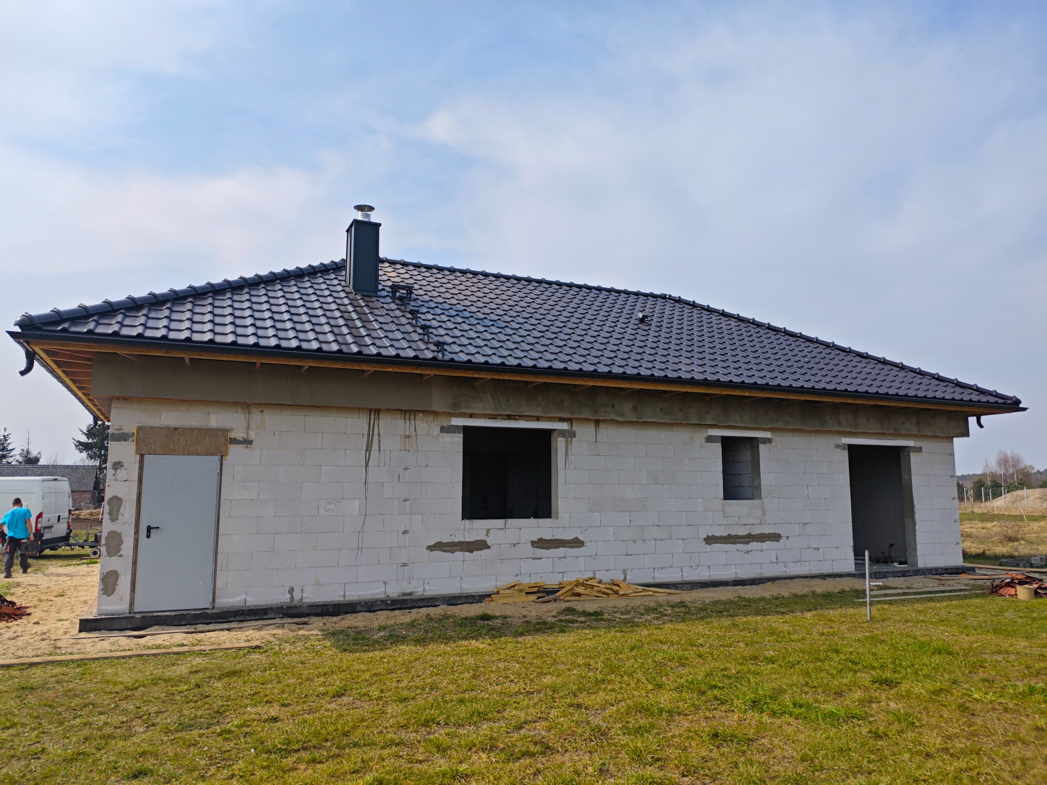 Dom w trakcie budowy z czarnym dachem z dachówki ceramicznej i widocznym kominem. Budynek z białych bloczków, okna i drzwi bez stolarki.