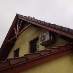 ProklimaTech Mariusz Stawarz - Zewnętrzna jednostka klimatyzacji zamontowana na ścianie domu z żółtą elewacją i brązowym dachem, widoczna z dołu pod kątem.