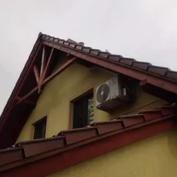 Zewnętrzna jednostka klimatyzacji zamontowana na ścianie domu z żółtą elewacją i brązowym dachem, widoczna z dołu pod kątem.