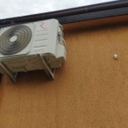 ProklimaTech Mariusz Stawarz - Zewnętrzna jednostka klimatyzatora zamontowana na pomarańczowej ścianie budynku, widoczny fragment rynny i pochmurne niebo w tle.