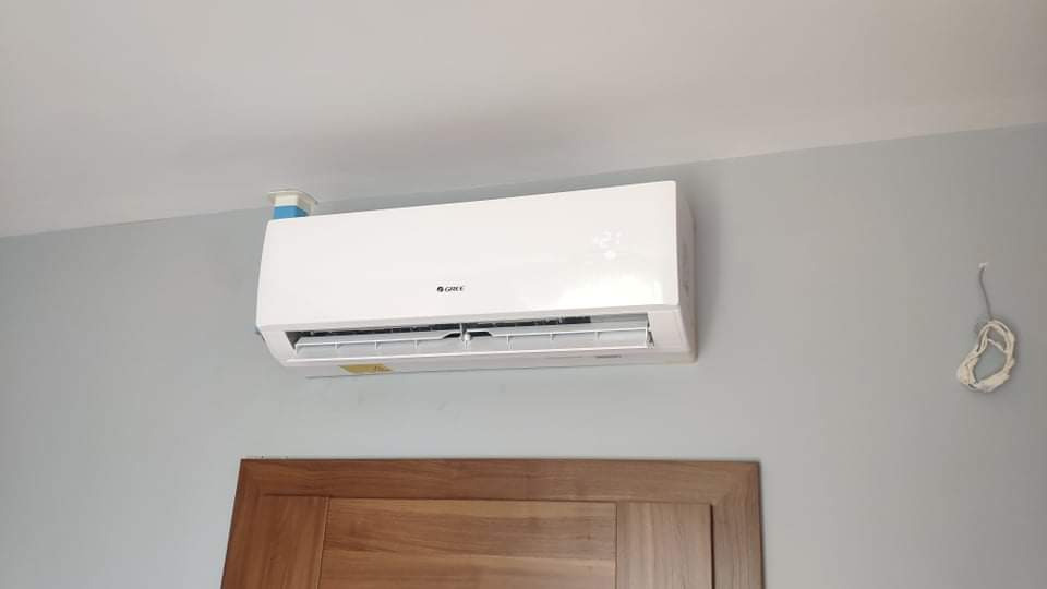 Biały klimatyzator marki Gree zamontowany na szarej ścianie nad drewnianymi drzwiami, widoczne wystające kable po prawej stronie.