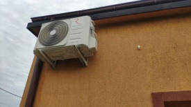 Zewnętrzna jednostka klimatyzatora zamontowana na pomarańczowej ścianie budynku, widoczny fragment rynny i pochmurne niebo w tle.