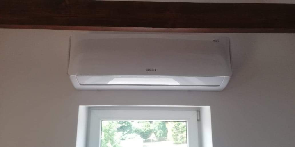 Biała jednostka wewnętrzna klimatyzatora zamontowana nad oknem w pokoju z drewnianą belką stropową.