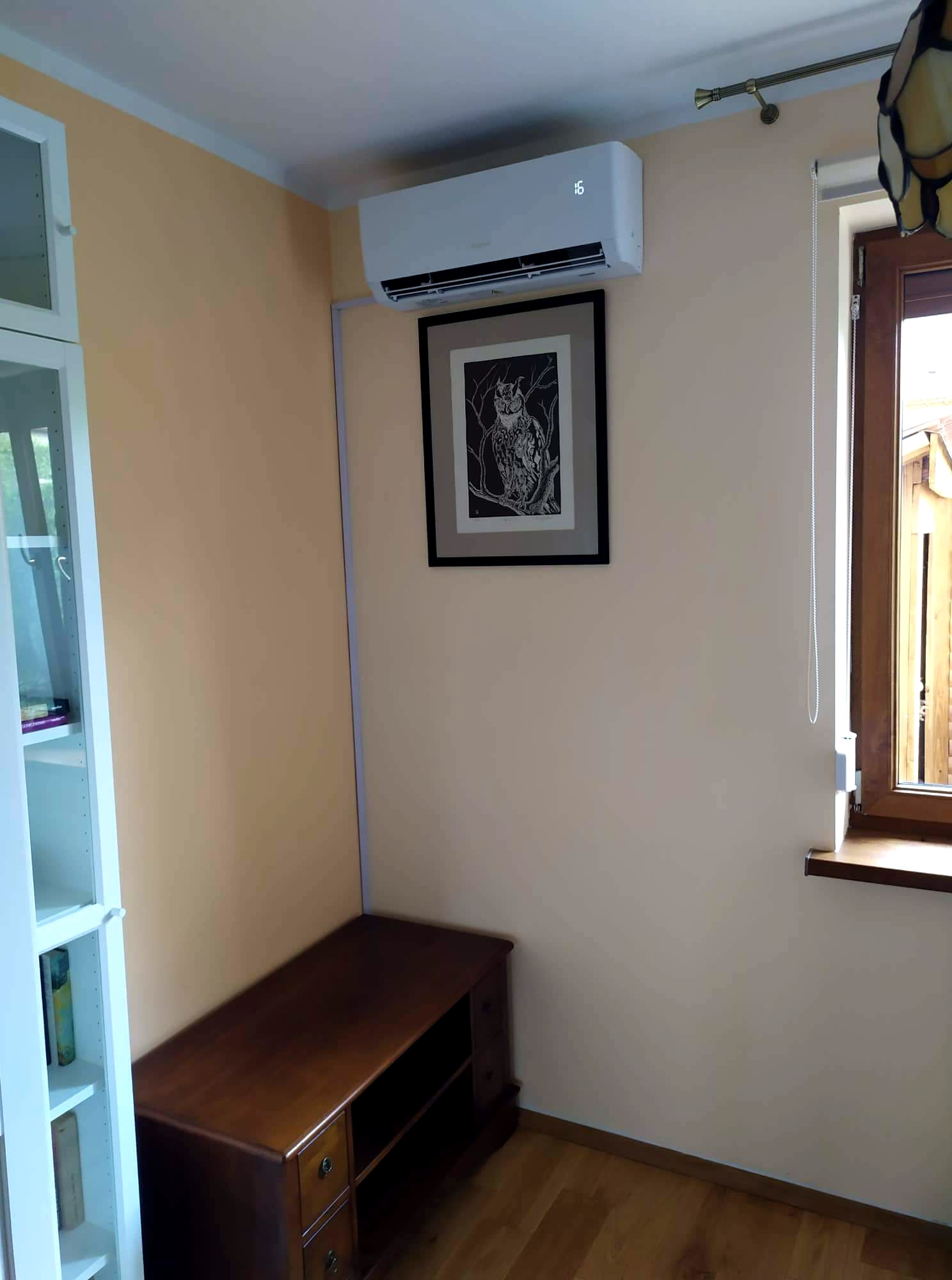 Biała jednostka klimatyzacyjna ścienna w pokoju z drewnianą podłogą, wisząca nad ciemnobrązową komodą, obok okna z drewnianą ramą i roletą. Na ścianie wisi oprawiony obraz z motywem sowy.