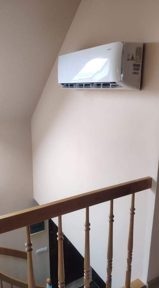 Biały klimatyzator ścienny zamontowany w rogu pokoju na poddaszu, widoczny fragment drewnianej balustrady schodów.