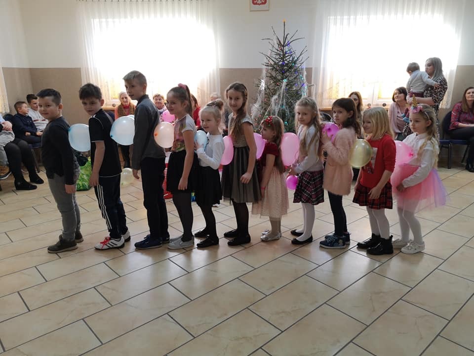 Dzieci ustawione w rzędzie trzymają balony, przygotowane do zabawy lub konkursu, w tle choinka i publiczność.